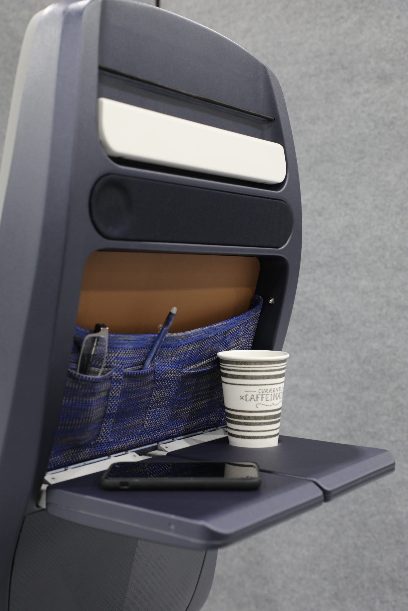 An airplane seat back with a folded tray table holding a paper cup and a smartphone. The seat pocket contains a tablet and a pen. The background is a plain gray surface.