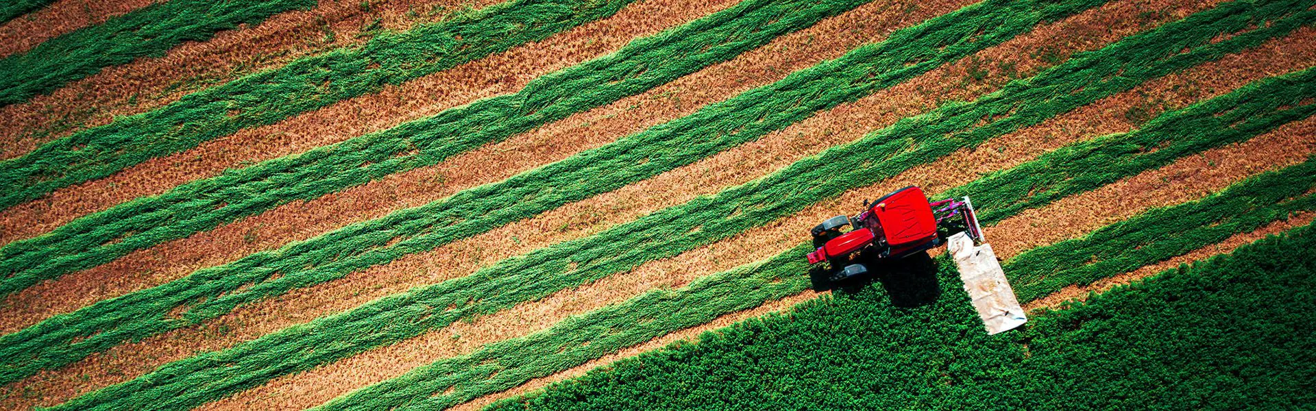 Aerial view of a red tractor harvesting a field, creating alternating green and brown stripes. The tractor moves diagonally across the farm landscape.