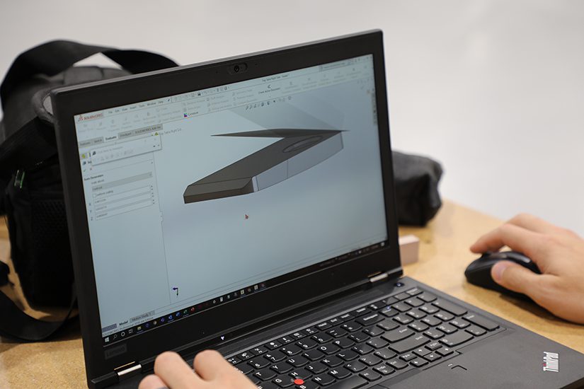 A person is using a laptop to design a 3D model of a sleek, flat object with angled edges, displayed on the screen. The workspace includes a mouse and a backpack in the background.