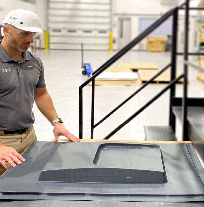 Man in a gray shirt and white hard hat examines a large, flat, rectangular object on a table in an industrial setting. Stairs and blurred equipment are visible in the background.