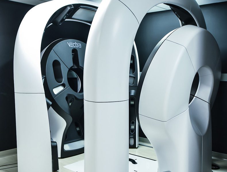 A futuristic room containing large, sleek grey machines with curved, circular designs and visible electronic components marked "Vectra." The machines are positioned closely together, creating an advanced technological atmosphere.