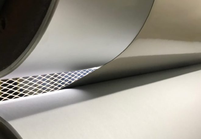 Close-up view of a large paper roll being processed, with a glimpse of a textured metal mesh visible between layers. The paper is smooth and bright, partially illuminated by light shining through the mesh design.