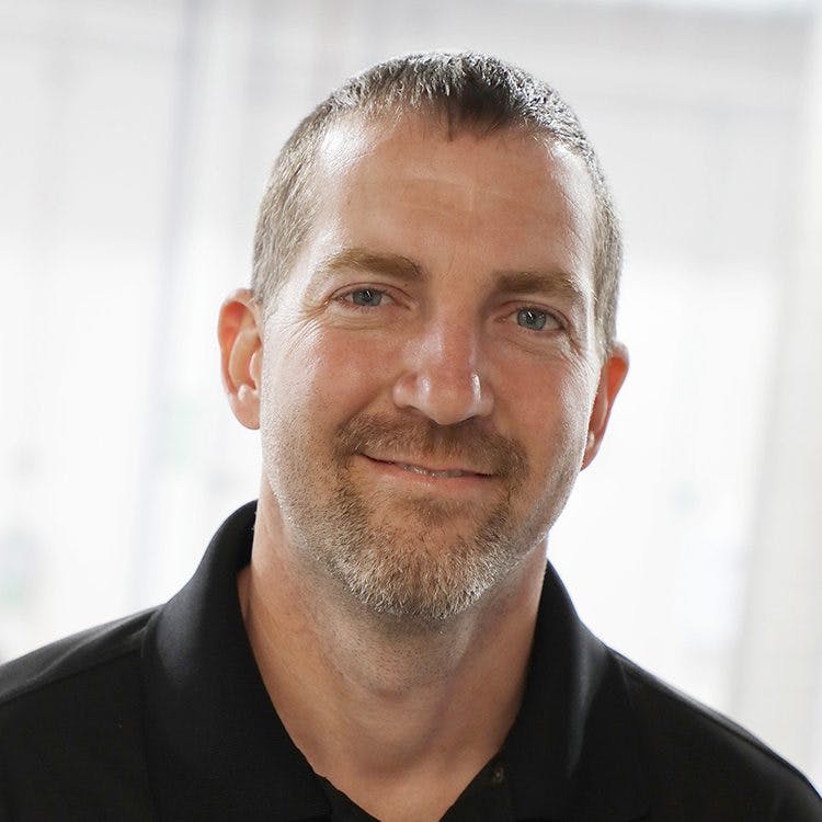 A man with short hair and a beard smiles slightly, wearing a black polo shirt. The background is softly blurred, suggesting an indoor setting with natural light.