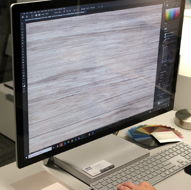 A person is using a large computer monitor displaying editing software, working on a digital wood texture. A keyboard, mouse, color swatches, and a note card are placed on the desk.