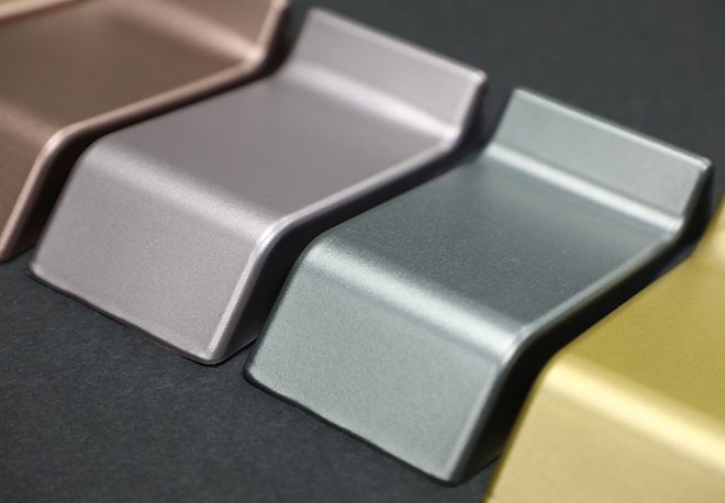 Close-up of four metallic paint samples in brown, gray, green, and yellow shades, placed side by side on a dark background. Each sample has a sleek, angled shape with a glossy finish, highlighting the colors' reflective qualities.