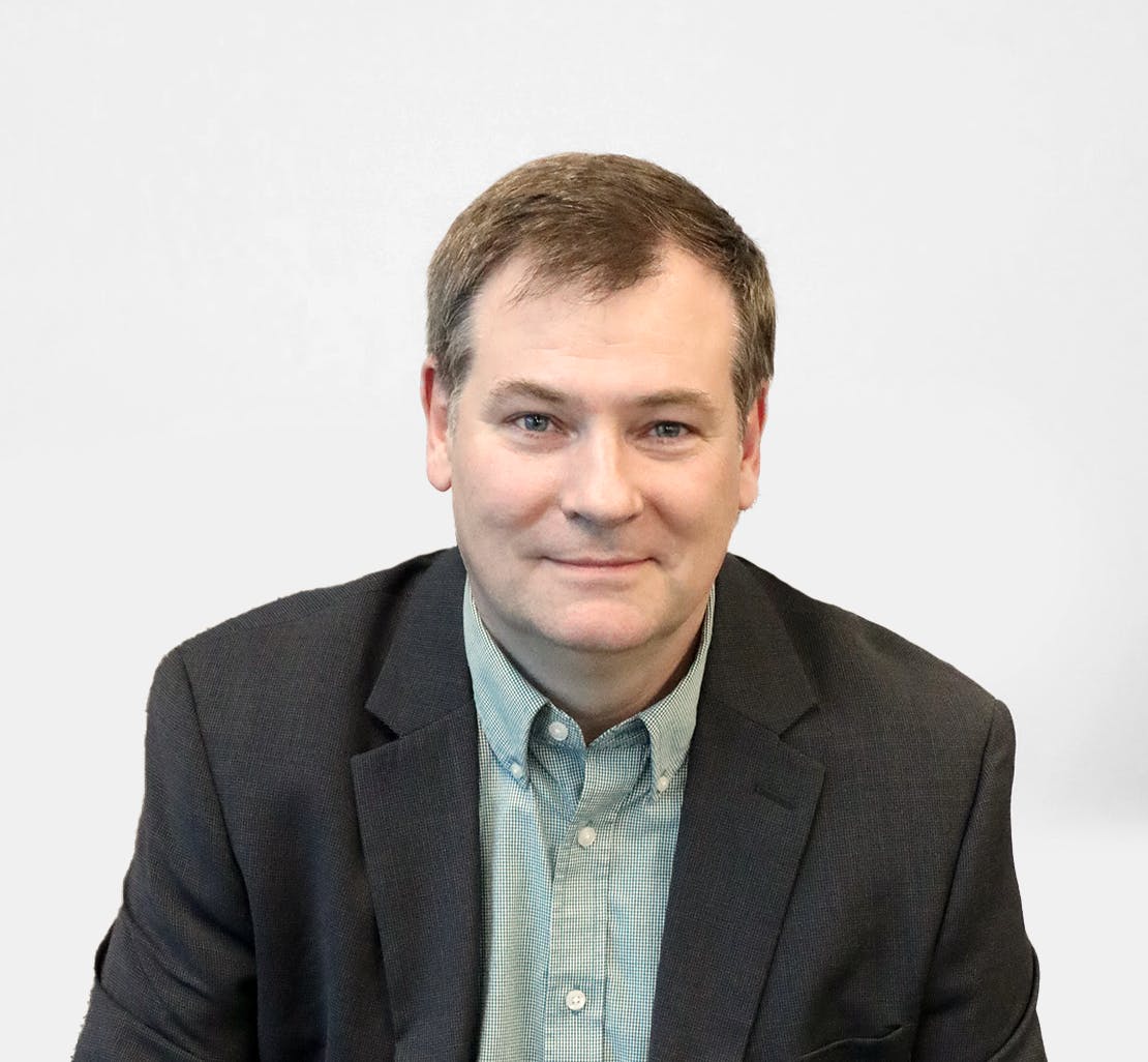 A man in a dark suit and light green shirt is looking at the camera with a neutral expression. The background is plain white.