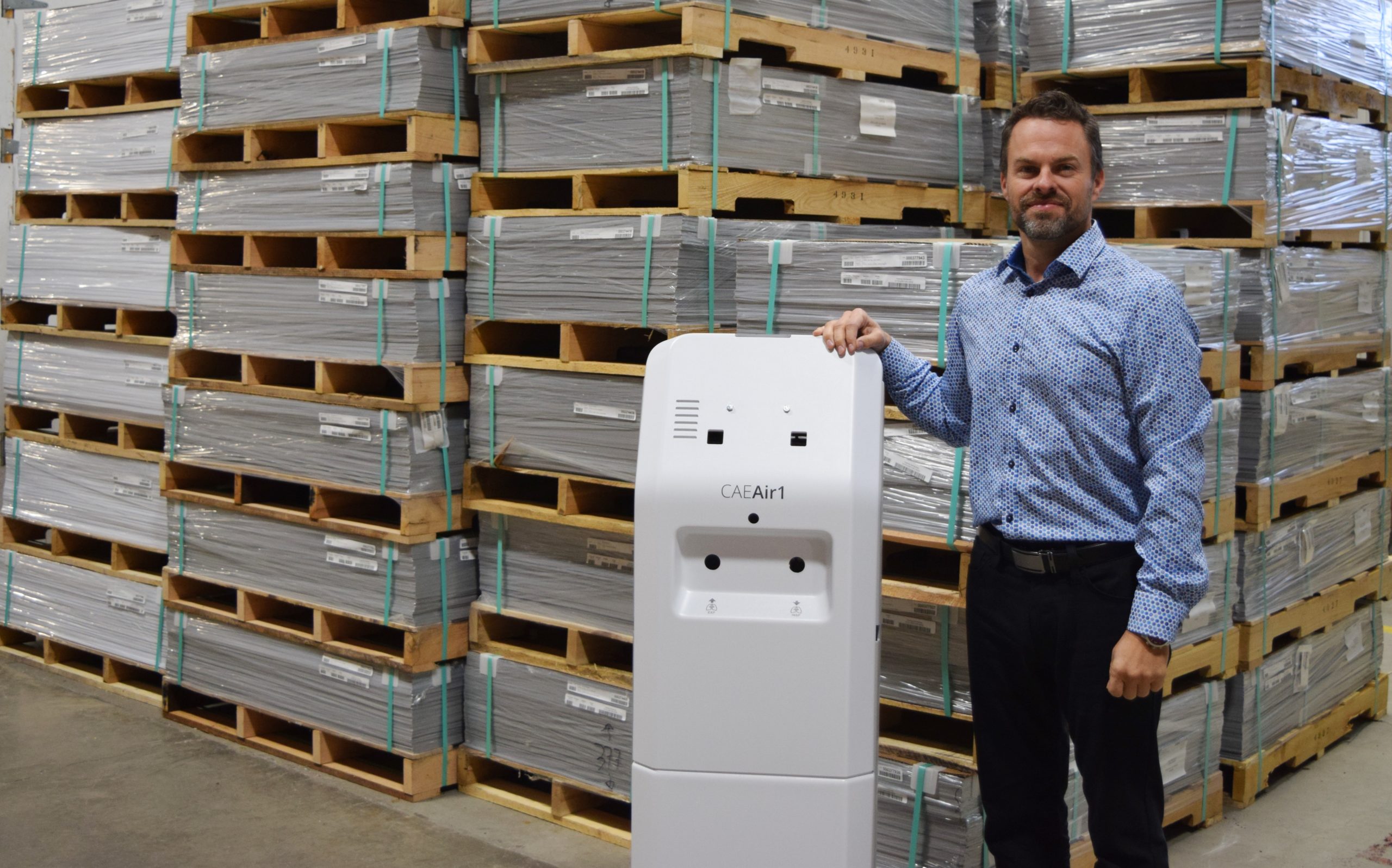 A person stands next to a tall, white air purification device labeled "CleaAir1" in a warehouse filled with stacked metal pallets. The individual is wearing a light blue patterned shirt and smiles at the camera.
