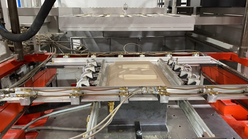 A machine with red and silver components is processing a flat rectangular object in a manufacturing setting. Numerous cables and tubes are connected, with a large metal plate visible in the background.