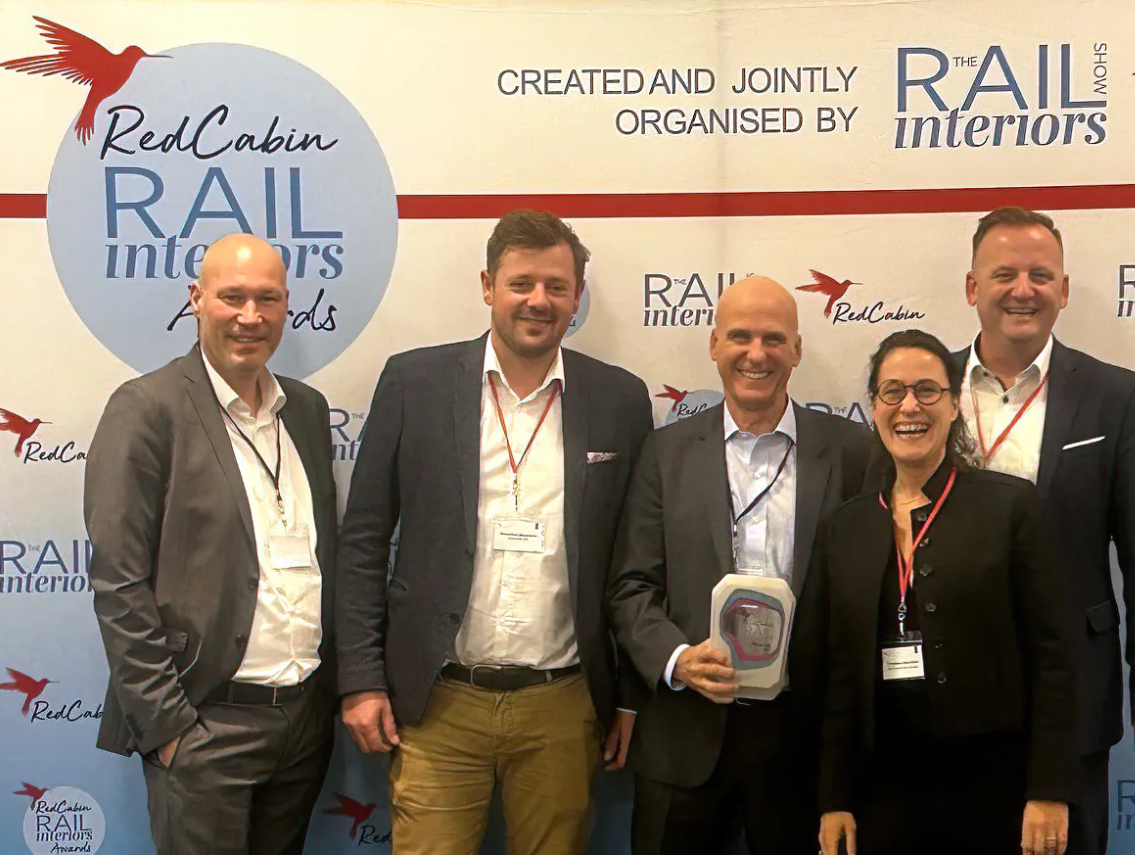 A group of five people, four men and one woman, are standing in front of a backdrop with logos for RedCabin Rail Interiors Awards. The man in the center is holding a trophy and all are smiling. They appear to be at an event.