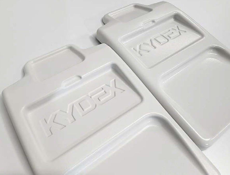 Two white rectangular KYDEX sheets with raised edges and a handle-like section at one end. The brand name "KYDEX" is embossed in the center of each sheet. They are placed on a smooth, light-colored surface.