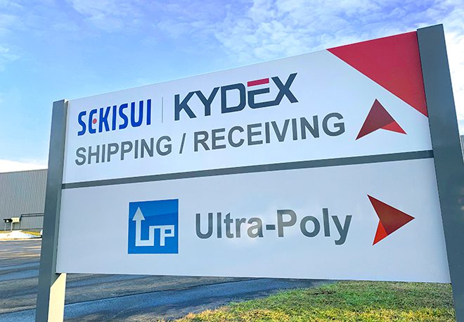 A sign with "SEKISUI KYDEX" and "Ultra-Poly" logos directs to "Shipping / Receiving." Two red arrows point right. The background features a cloudy sky and a partial view of a building.